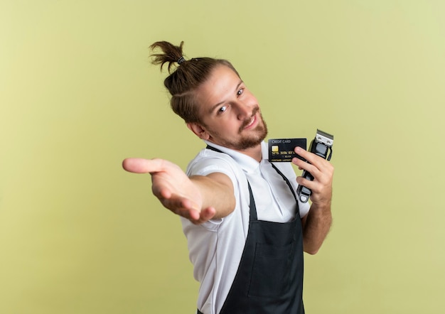 Erfreulicher junger hübscher Friseur, der Kreditkarte und Haarschneidemaschine hält und Hand an der Front lokalisiert auf olivgrüner Wand ausstreckt