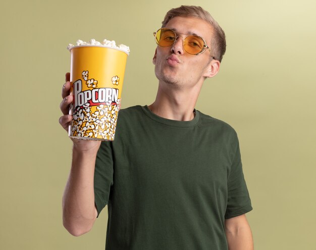 Erfreulicher junger hübscher Kerl, der grünes Hemd mit Gläsern hält, die Eimer Popcorn halten, die Kussgeste lokalisiert auf olivgrüner Wand zeigt