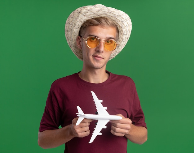 Erfreulicher junger hübscher kerl, der rotes hemd mit brille und hut hält spielzeugflugzeug lokalisiert auf grüner wand trägt