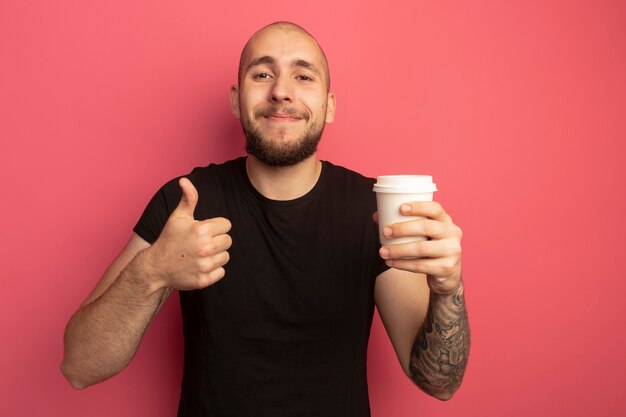 Erfreulicher junger hübscher Kerl, der Tasse Kaffee hält und Daumen zeigt