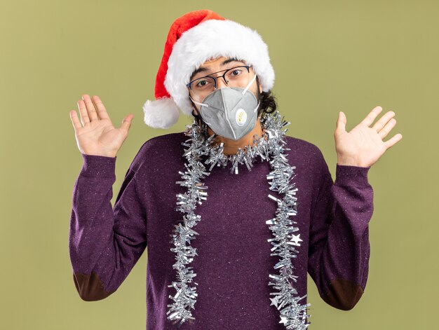 Erfreulicher junger hübscher Kerl, der Weihnachtshut und medizinische Maske mit Girlande am Hals trägt, die Hände lokalisiert auf olivgrünem Hintergrund verbreiten