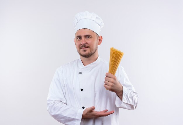 Erfreulicher junger hübscher Koch in der Kochuniform, die auf Spaghetti-Nudeln hält und auf Weißraum lokalisiert zeigt