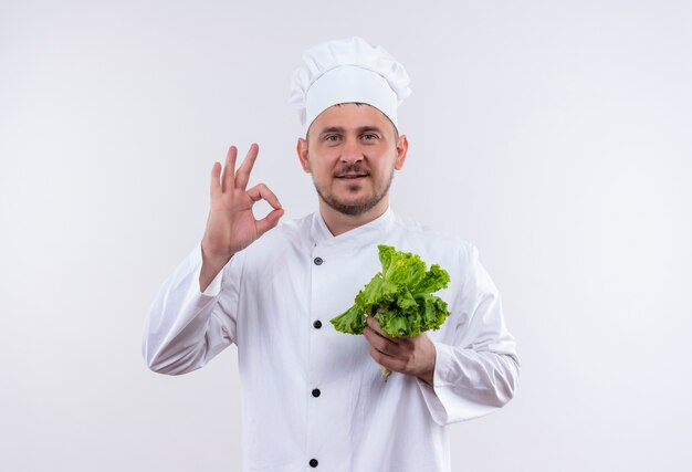 Erfreulicher junger hübscher Koch in der Kochuniform, die Salat hält und ok Zeichen lokalisiert auf weißem Raum tut