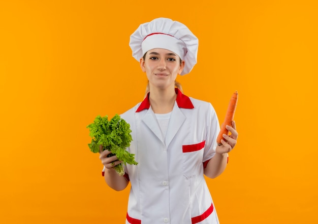 Erfreulicher junger hübscher Koch in der Kochuniform, die Salat und Karotte lokalisiert auf Orangenraum hält