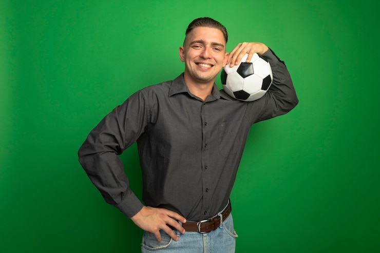 Kostenlose Foto erfreulicher junger hübscher mann im grauen hemd, der fußball auf seiner schulter hält, die zuversichtlich über grüner wand stehend lächelt