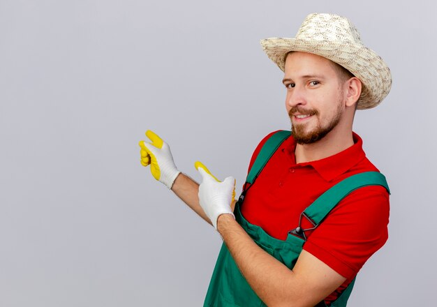 Erfreulicher junger hübscher slawischer Gärtner in der Uniform, die Gartenhandschuhe und Hut trägt, die in der Profilansicht stehen und auf die Seite zeigen, die lokalisiert zeigt