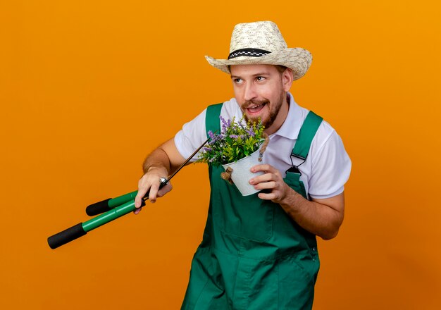 Erfreulicher junger hübscher slawischer Gärtner in der Uniform und im Hut, die Blumenpflanze und Gartenscheren halten, die lokal suchen