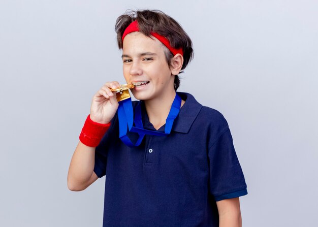 Erfreulicher junger hübscher sportlicher Junge, der Stirnband und Armbänder mit Zahnspangen und Medaille um den Hals trägt und die seitliche Beißmedaille lokalisiert auf weißer Wand betrachtet
