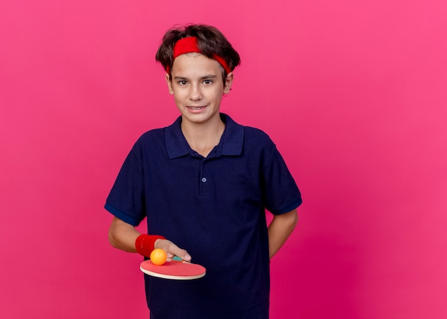 Erfreulicher junger hübscher sportlicher Junge, der Stirnband und Armbänder trägt, Tischtennisschläger mit Ball darauf hält und Hand hinter dem Rücken isoliert hält