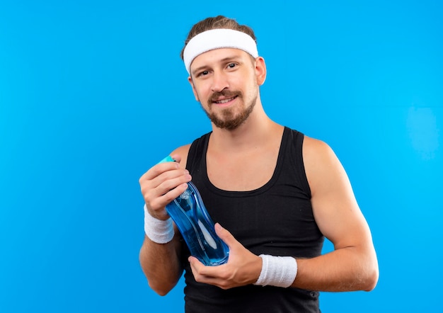 Erfreulicher junger hübscher sportlicher Mann, der Stirnband und Armbänder hält Wasserflasche hält, die auf blauem Raum lokalisiert