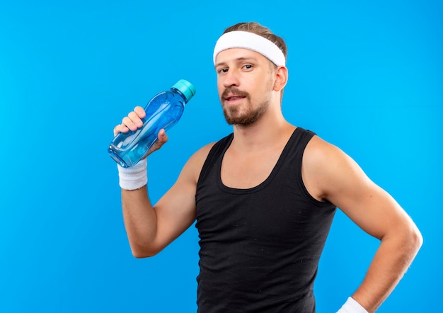 Erfreulicher junger hübscher sportlicher Mann, der Stirnband und Armbänder hält Wasserflasche hält, die auf blauem Raum lokalisiert