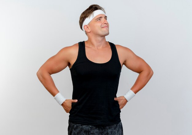 Erfreulicher junger hübscher sportlicher Mann, der Stirnband und Armbänder trägt, die Hände auf Taille setzen und Seite lokalisiert auf weißer Wand betrachten