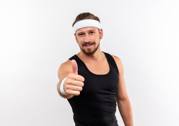 Erfreulicher junger hübscher sportlicher Mann, der Stirnband und Armbänder trägt, zeigt Daumen oben auf weißem Raum