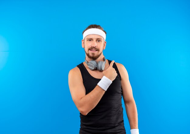 Erfreulicher junger hübscher sportlicher Mann, der Stirnband und Armbänder und Kopfhörer um den Hals trägt, der auf Seite zeigt, die auf blauem Raum isoliert ist