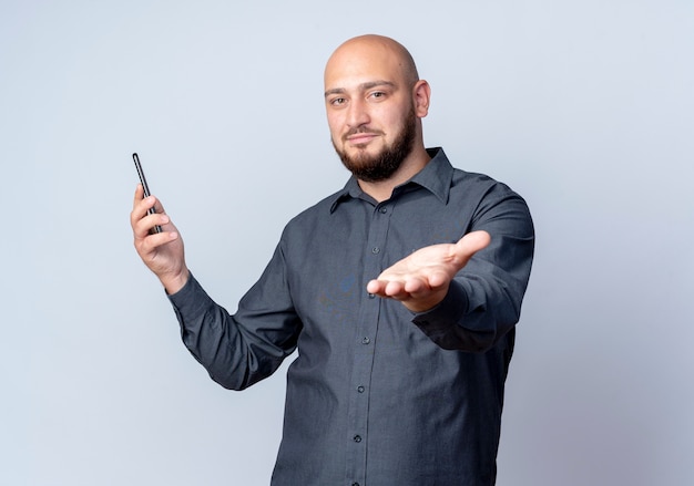Erfreulicher junger kahlköpfiger Callcenter-Mann, der Handy hält und Hand an Kamera lokalisiert auf weißem Hintergrund mit Kopienraum ausdehnt