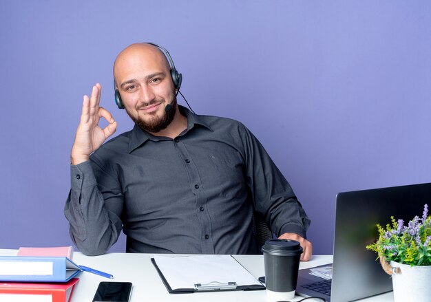 Erfreulicher junger kahlköpfiger Callcenter-Mann, der Headset trägt, das am Schreibtisch mit Arbeitswerkzeugen sitzt, die ok Zeichen lokalisiert auf lila Hintergrund tun