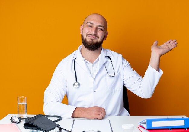 Erfreulicher junger kahlköpfiger männlicher Arzt, der medizinische Robe und Stethoskop trägt, die am Schreibtisch mit medizinischen Werkzeugpunkten mit Hand an der Seite lokalisiert auf orange Hintergrund sitzen