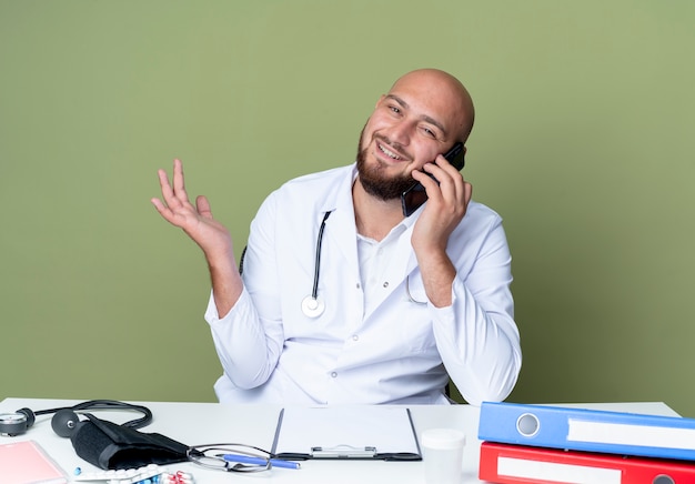 Erfreulicher junger kahlköpfiger männlicher Arzt, der medizinische Robe und Stethoskop trägt, die an Schreibtischarbeit mit medizinischen Werkzeugen sitzen, spricht am Telefon lokalisiert auf grünem Hintergrund