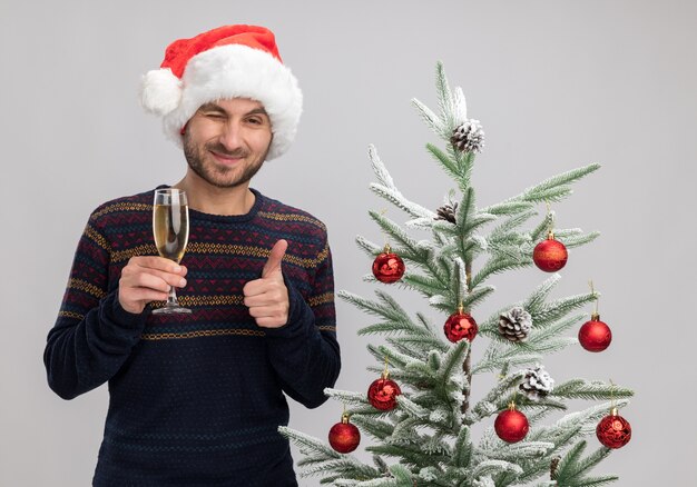 Erfreulicher junger kaukasischer Mann, der Weihnachtshut trägt, der nahe Weihnachtsbaum hält, der Glas Champagner hält, der Kamera betrachtet, die zwinkert und Daumen oben auf weißem Hintergrund zeigt