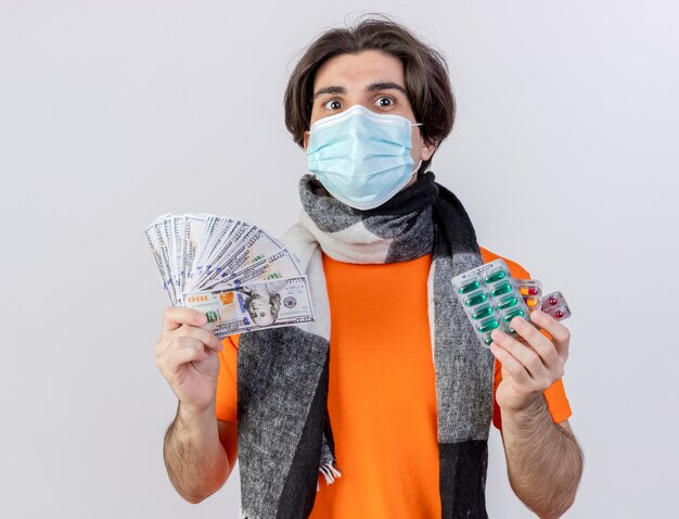 Erfreulicher junger kranker Mann, der Schal und medizinische Maske hält, die Geld mit Pillen lokalisiert auf weißem Hintergrund hält