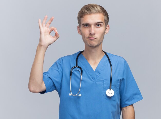 Erfreulicher junger männlicher Arzt, der Arztuniform mit Stethoskop trägt, das auf weiße Wand lokalisierte gute Geste zeigt