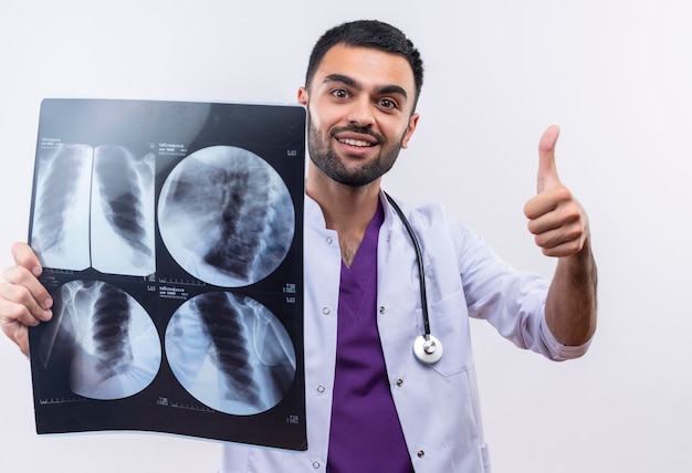 Erfreulicher junger männlicher Arzt, der das medizinische Kleid des Stethoskops trägt, das Röntgen seinen Daumen oben auf lokalisiertem weißem Hintergrund hält