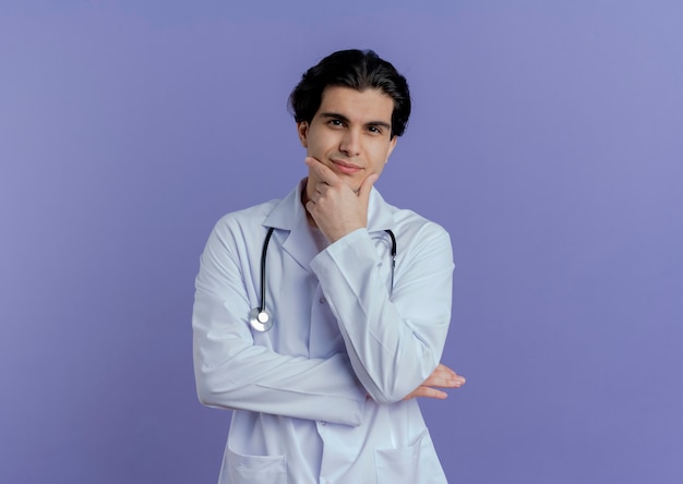 Erfreulicher junger männlicher Arzt, der medizinische Robe und Stethoskop trägt, die Hand auf Kinn lokalisiert auf lila Wand mit Kopienraum setzen