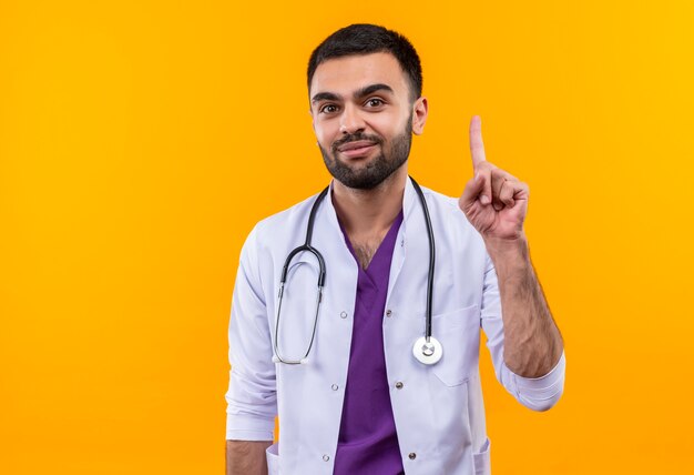 Erfreulicher junger männlicher Arzt, der medizinisches Kleid des Stethoskops trägt, zeigt auf lokalisierten gelben Hintergrund