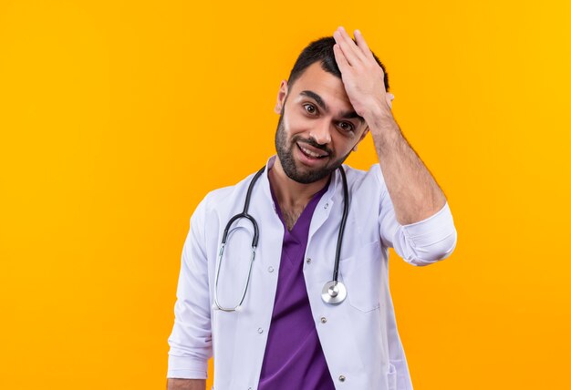 Erfreulicher junger männlicher Arzt, der Stethoskop-medizinisches Kleid trägt, legte seine Hand auf Kopf auf lokalisiertem gelbem Hintergrund