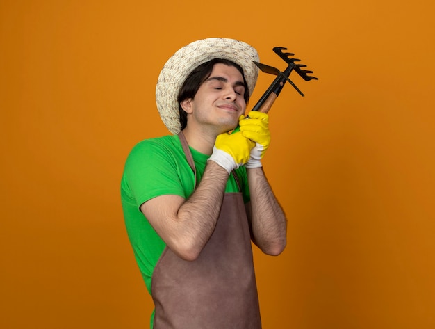 Erfreulicher junger männlicher Gärtner in der Uniform, die Gartenhut mit Handschuhen mit geschlossenen Augen trägt, die Rechen mit Hackrechen um Gesicht lokalisiert auf Orange halten