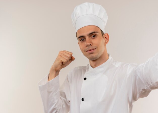 Erfreulicher junger männlicher Koch, der Kochuniform hält Kamera hält, die starke Geste zeigt
