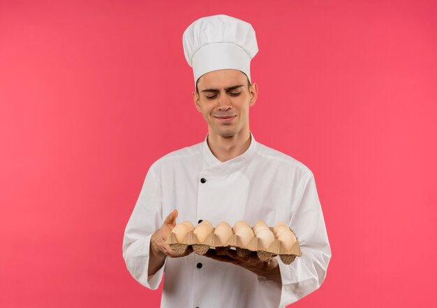 Erfreulicher junger männlicher Koch, der Kochuniform trägt und Charge der Eier in seiner Hand betrachtet