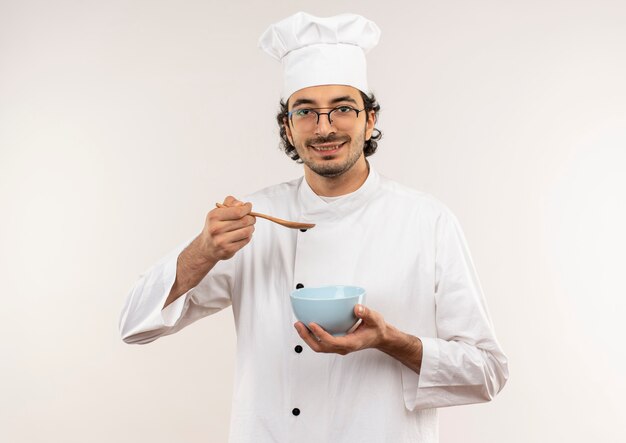 Erfreulicher junger männlicher Koch, der Kochuniform und Gläser hält Löffel mit Schüssel isoliert auf weißer Wand trägt