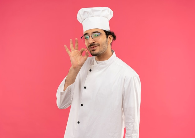 Erfreulicher junger männlicher Koch, der Kochuniform und Gläser trägt, die köstliche Geste lokalisiert auf rosa Wand zeigen