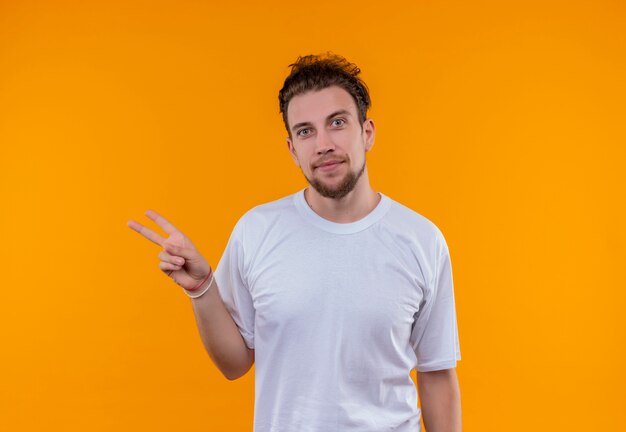 Erfreulicher junger Mann, der weißes T-Shirt trägt, das Friedensgeste auf lokalisiertem orange Hintergrund zeigt