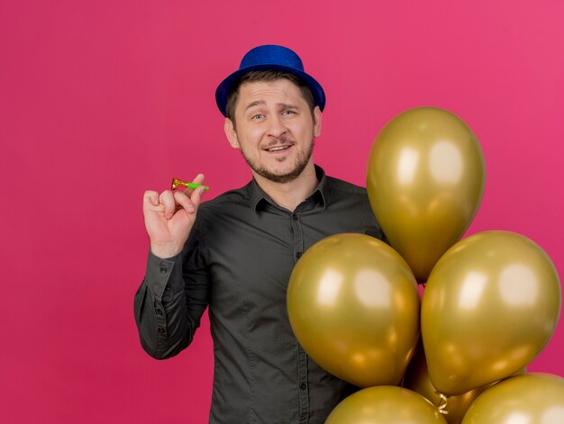 Erfreulicher junger Party-Typ, der blauen Hut hält, der Luftballons mit Partygebläse hält, die auf Rosa lokalisiert werden