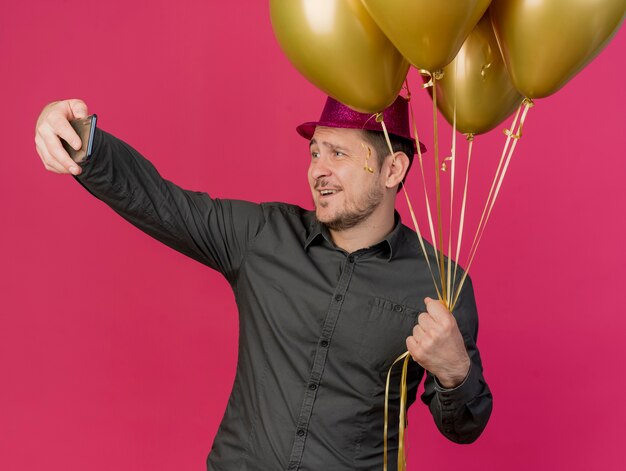 Erfreulicher junger Party-Typ, der rosa Hut hält, der Luftballons hält, nehmen ein Selfie, das auf rosa isoliert wird