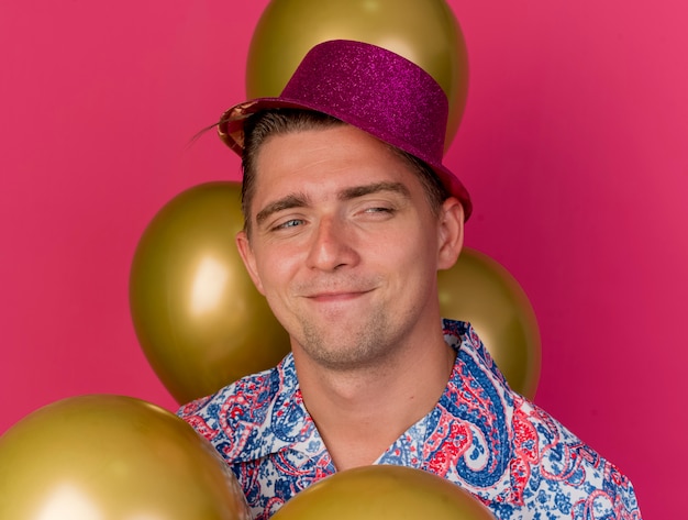 Erfreulicher junger Party-Typ, der rosa Hut trägt, der unter Luftballons steht, die auf rosa lokalisiert werden