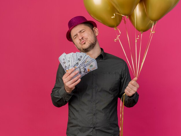 Erfreulicher junger Party-Typ mit geschlossenen Augen, der rosa Hut trägt, der Bargeld mit Luftballons hält, die auf rosa isoliert werden