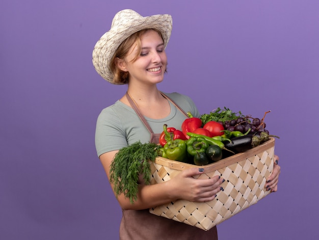 Erfreulicher junger slawischer weiblicher Gärtner, der Gartenhut hält und Gemüsekorb auf lila betrachtet