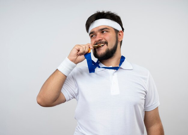 Erfreulicher junger sportlicher Mann, der Seite betrachtet, die Stirnband und Armband trägt, und Bissmedaille lokalisiert auf weißer Wand