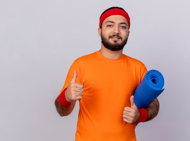 Erfreulicher junger sportlicher Mann, der Stirnband und Armband hält, die Yogamatte halten