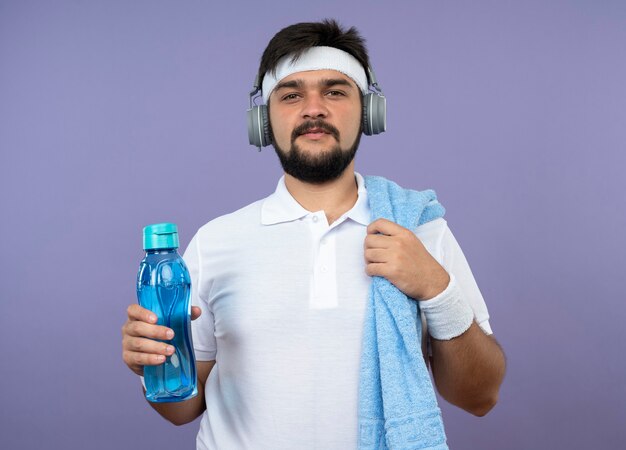 Erfreulicher junger sportlicher Mann, der Stirnband und Armband mit Kopfhörern hält, die Wasserflasche mit Handtuch auf Schulter halten