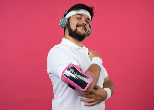 Erfreulicher junger sportlicher Mann, der Stirnband und Armband mit Kopfhörern und Telefonarmband trägt, lokalisiert auf rosa Wand