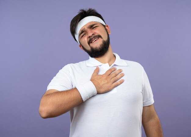 Erfreulicher junger sportlicher Mann, der Stirnband und Armband trägt und Hand auf Brust legt