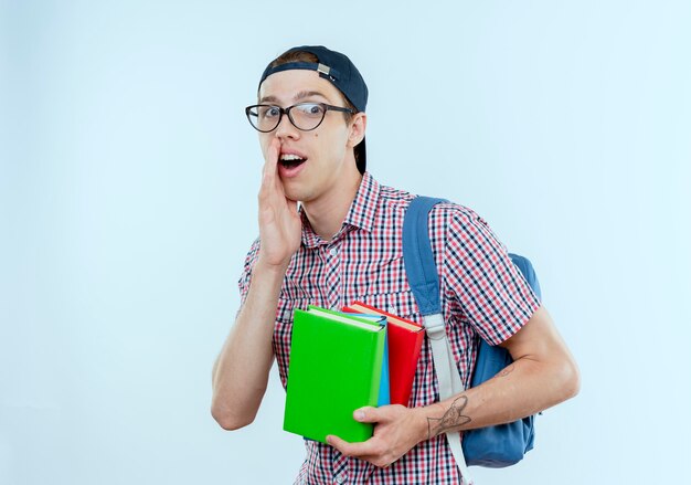Erfreulicher junger Studentjunge, der Rückentasche und Brille und Kappe trägt Bücher und Flüstern auf Weiß hält
