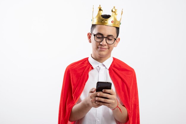 Erfreulicher junger Superheldenjunge im roten Umhang, der Brille und Krone unter Verwendung des Mobiltelefons trägt, das auf weißem Hintergrund mit Kopienraum lokalisiert wird