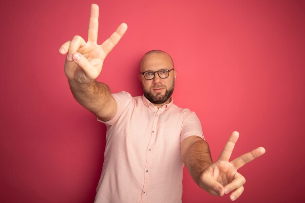 Erfreulicher kahler Mann mittleren Alters, der rosa T-Shirt mit Brille zeigt, die Friedensgeste lokalisiert auf Rosa zeigt