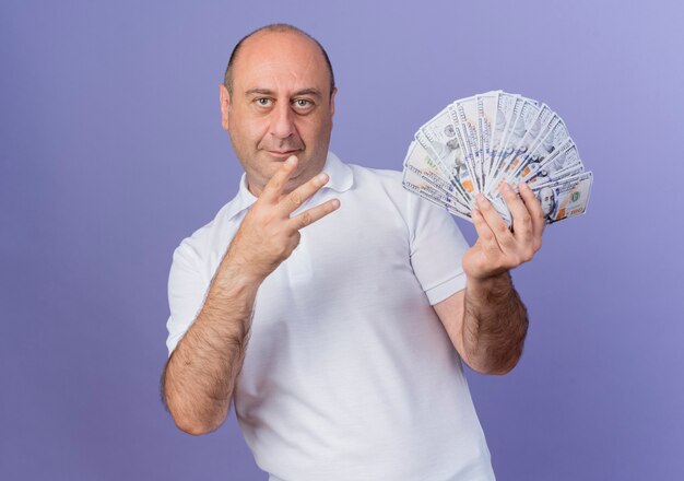 Erfreulicher lässiger reifer Geschäftsmann, der Geld hält, das Seite betrachtet und drei mit Hand lokalisiert auf lila Hintergrund mit Kopienraum zeigt