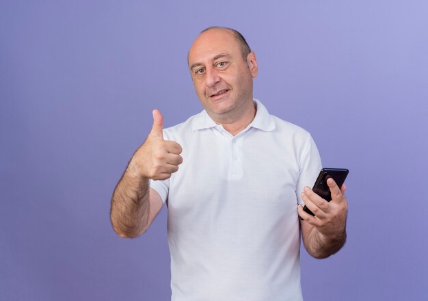 Erfreulicher lässiger reifer Geschäftsmann, der Kamera betrachtet, die Handy hält und Daumen oben auf lila Hintergrund mit Kopienraum zeigt